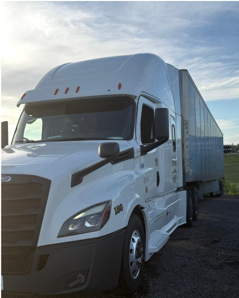 White Freightliner tractor with a trailer parked beside an open field at sunrise.