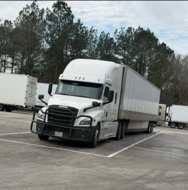 White Freightliner sleeper tractor with a trailer parked in a paved lot with trees in the background.