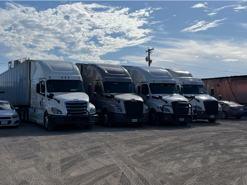 Four sleeper tractors parked side by side in a lot under a partly cloudy sky.